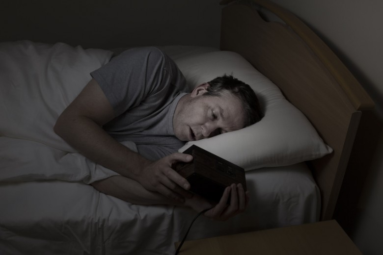 Mature man, eyes open with both hands on alarm clock while checking time, cannot sleep at night from insomnia